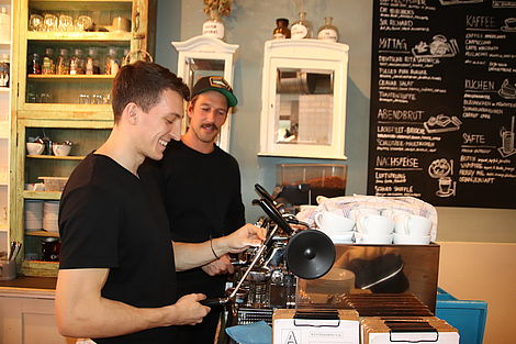 Genuss ist ihr Job: Zwei Männer stehen an einer Kaffeemaschine in einem Café.