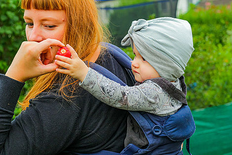 Eine Frau trägt ein Kind auf ihrem Rücken und reicht diesem eine Erdbeere.