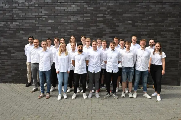 Eine Gruppe von zwanzig Studenten in weißen T-Shirts steht vor einer grauen Mauerwand. 