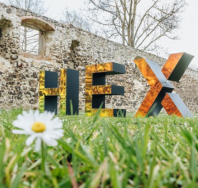 Auf einer Wiese sind vor den Mauern einer Ruine die Buchstaben HEX zu lesen. (Bildrechte: eventives)