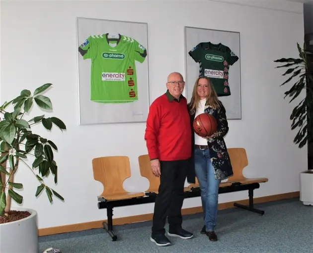 Ein glatzköpfiger mann im roten Pullover und eine Frau mit langen rötlichen Haaren stehen nebeneinander, sie hält einen Basketball.