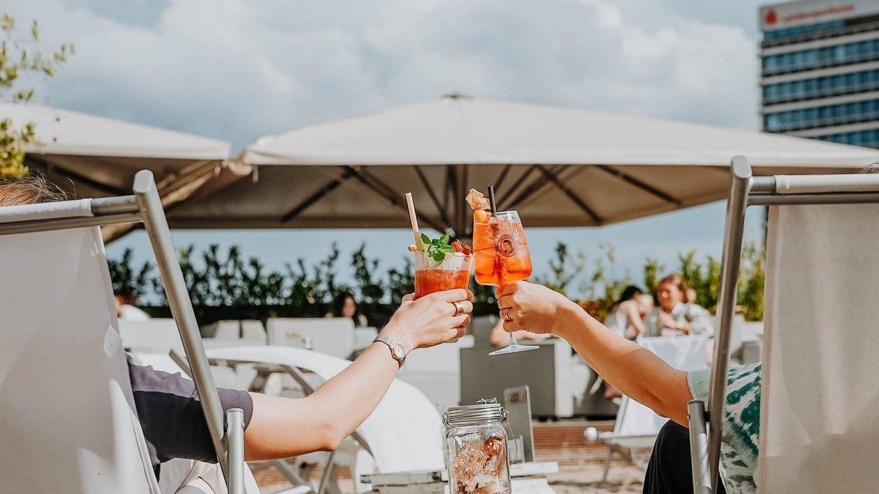 Zwei Menschen auf Liegestühlen in einer Beachbar, die mit einem Cocktail anstoßen 