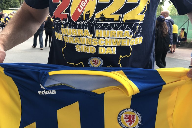 Ein Mann trägt ein buntes T-Shirt mit einer Gratulation zum Mannschaftsaufstieg und hält ein Trikot vor sich.