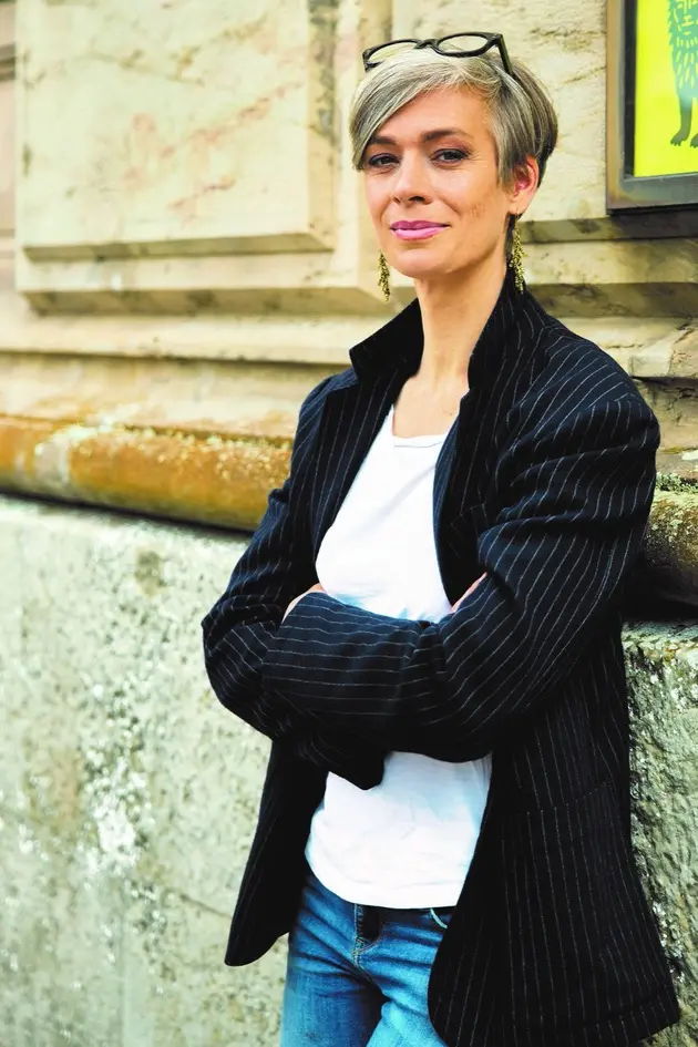 alt="Eine kurzhaarige Frau im Sako und T-Shirt lehnt an einer Marmorfassade."