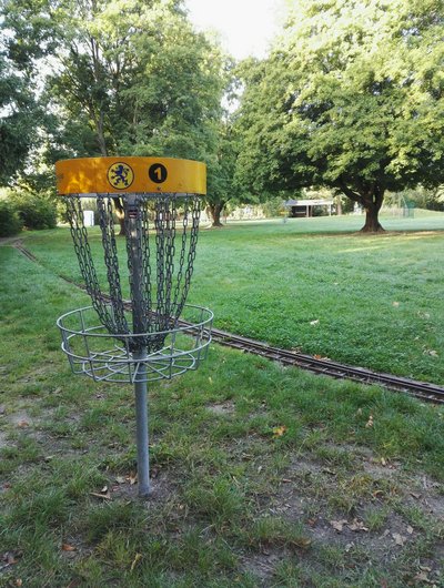 Man blickt auf einen Discgolfkorb auf einer Wiese in einer Parkanlage.