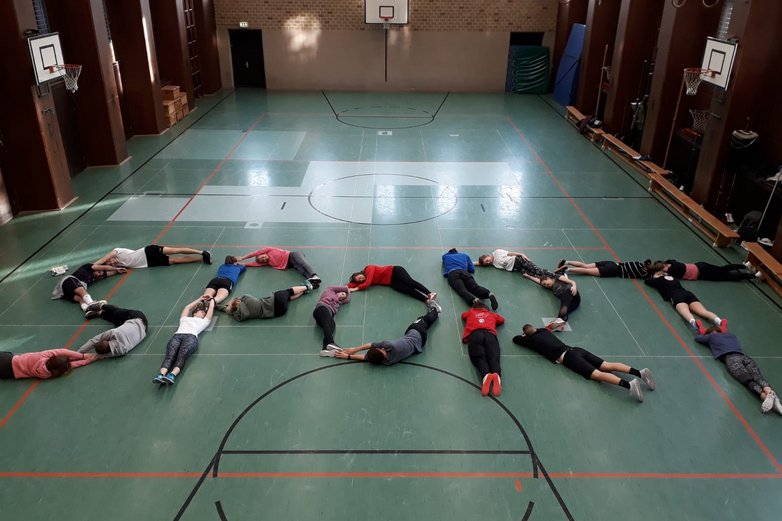 Junge Frauen und Männer formen auf dem Hallenboden mit ihren Körpern das Wort "Sport".