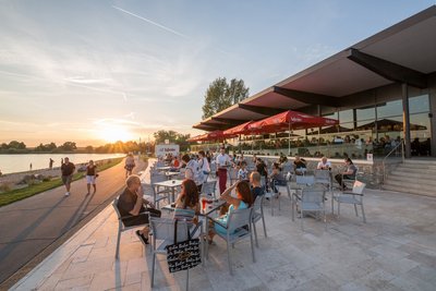 Man blickt auf die gefüllte Terrasse der Gastronomie am Salzgittersee. Am linken Bildrand sieht man den asphaltierten Fußweg und den See.