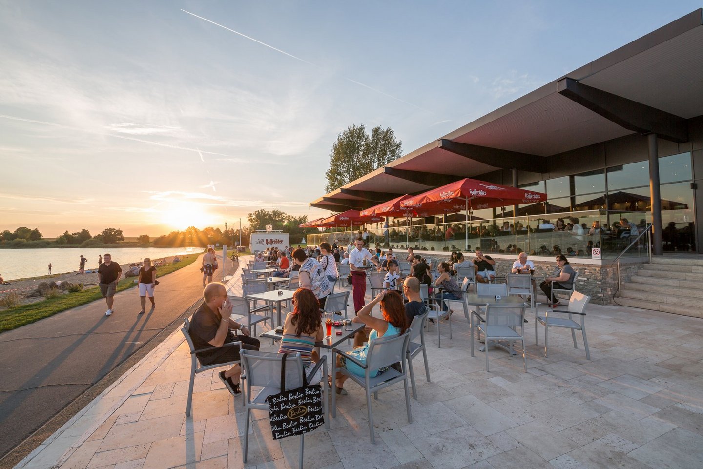 Man blickt auf die gefüllte Terrasse der Gastronomie am Salzgittersee. Am linken Bildrand sieht man den asphaltierten Fußweg und den See.