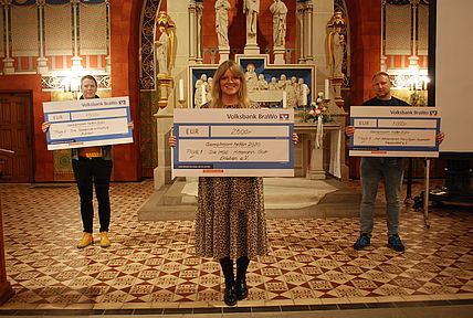 Zwei Frauen und ein Mann präsentieren große Gewinner-Schecks in einer Peiner Kirche. 