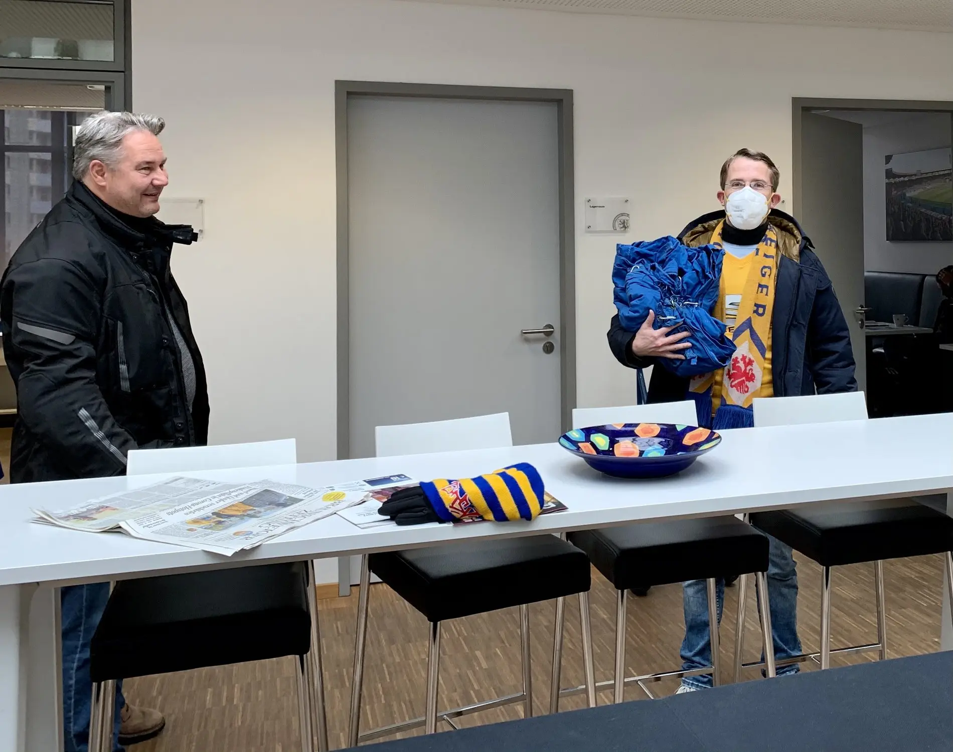 Zwei Männer mit Abstand und Maske in einem modernen Raum. einer davon in Eintracht-Trikot.