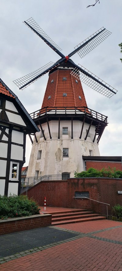 Man blickt auf eine Windmühle mit weißer Fassade und roten Dachziegeln. Auf mitte der Bauhöhe verläuft rings um die Windmühle ein Holzbalkon.