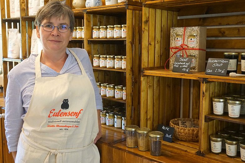 Eine Frau in Schürze vor einem Regal mit Senfgläsern.