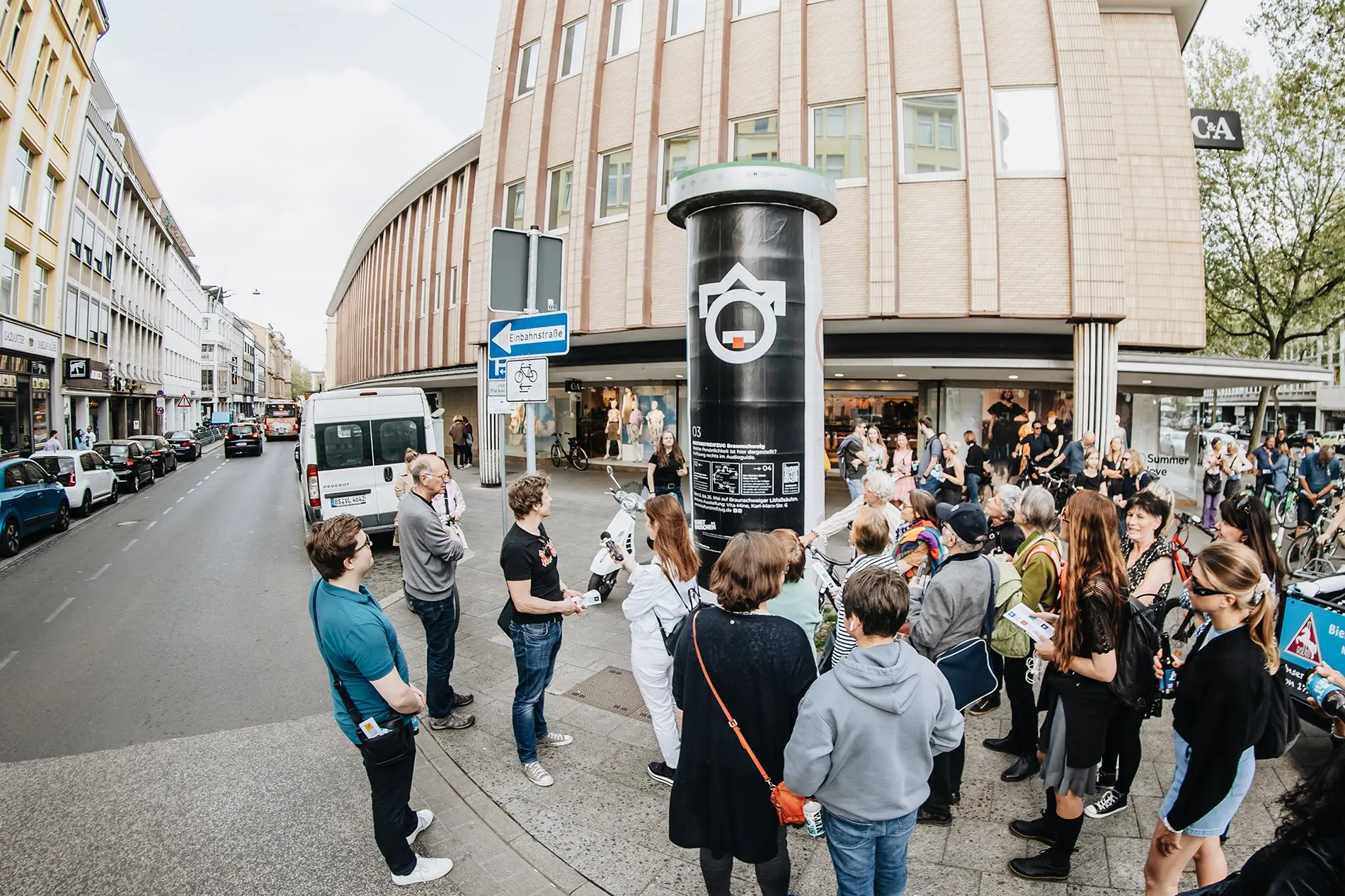 Eine Menschentraube steht vor einer Litfaßsäule.