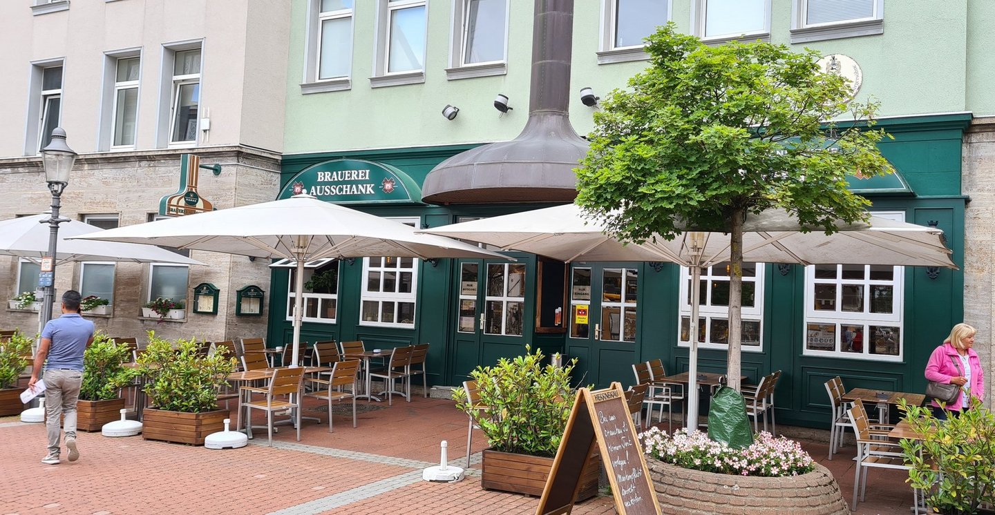 Man blickt auf eine Kneipe in der Peiner Innenstadt. Die Fassade des Gebäudes ist in dunkelgrün gehalten. Im Außenbereich in der Fußgängerzone sind Sitzplätze arrangiert.