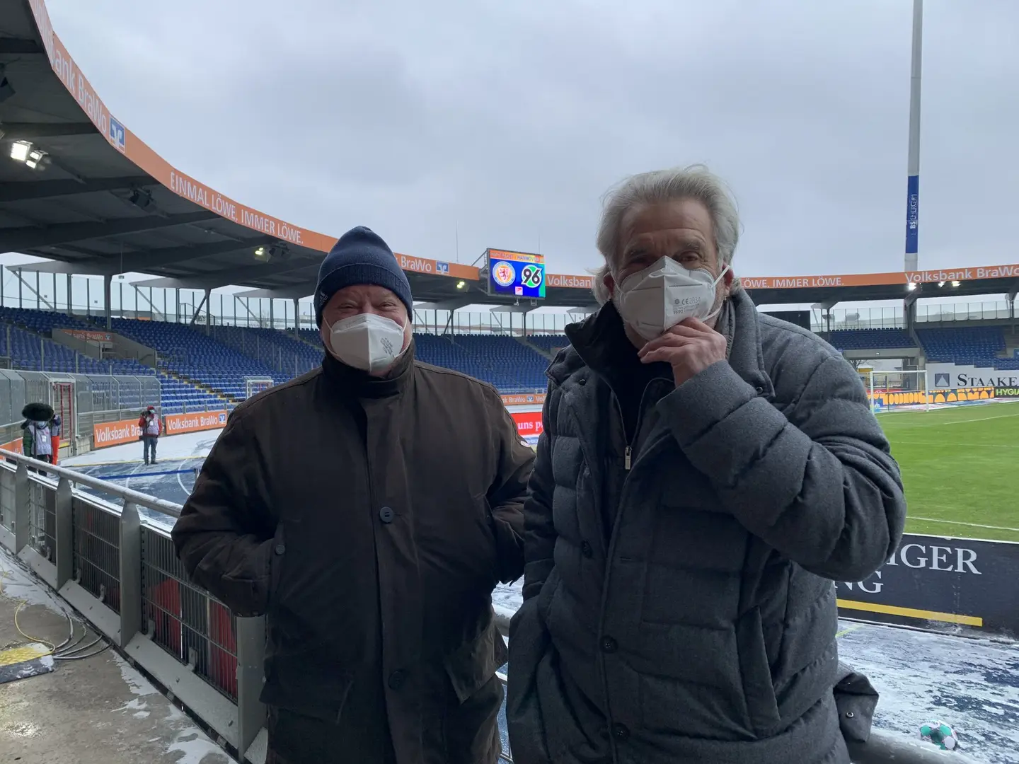 Zwei Männer mit Mundschutz stehen nebeneinander im Stadion.