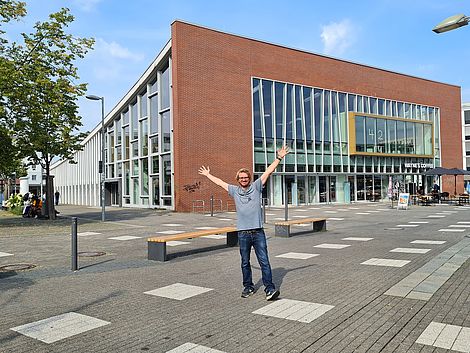 Ein Mann steht in jubelnder Geste vor einem modernen, kubusförmigen Gebäude.