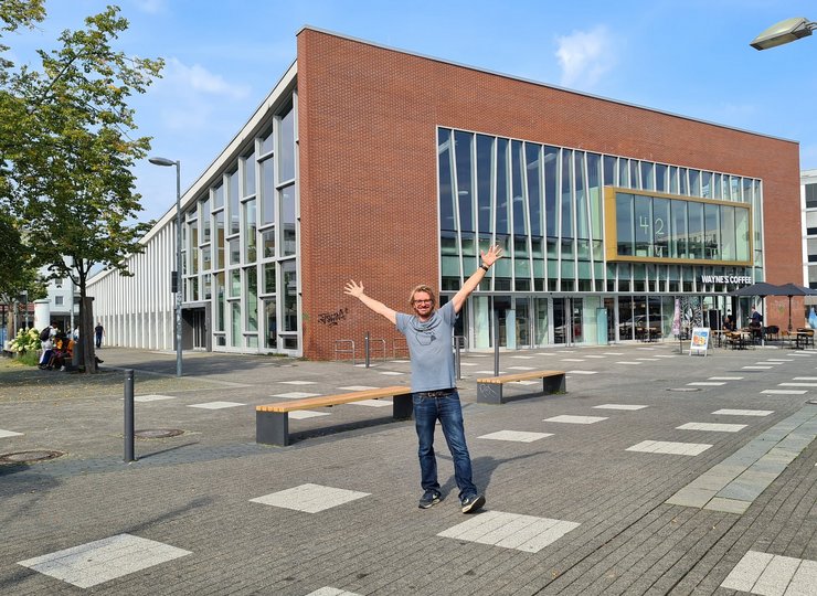 Ein Mann steht in jubelnder Geste vor einem modernen, kubusförmigen Gebäude.