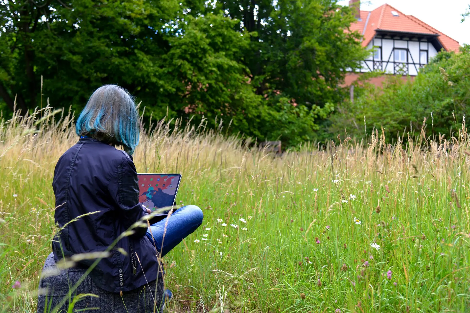 Eine Frau sitzt mit einem Laptop auf dem Schoß in einer Wiese.
