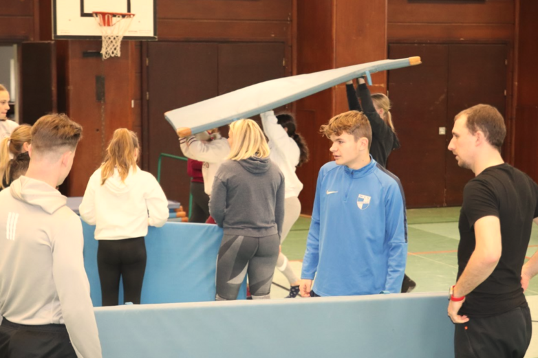 Junge Menschen mit Matten in einer Sporthalle.