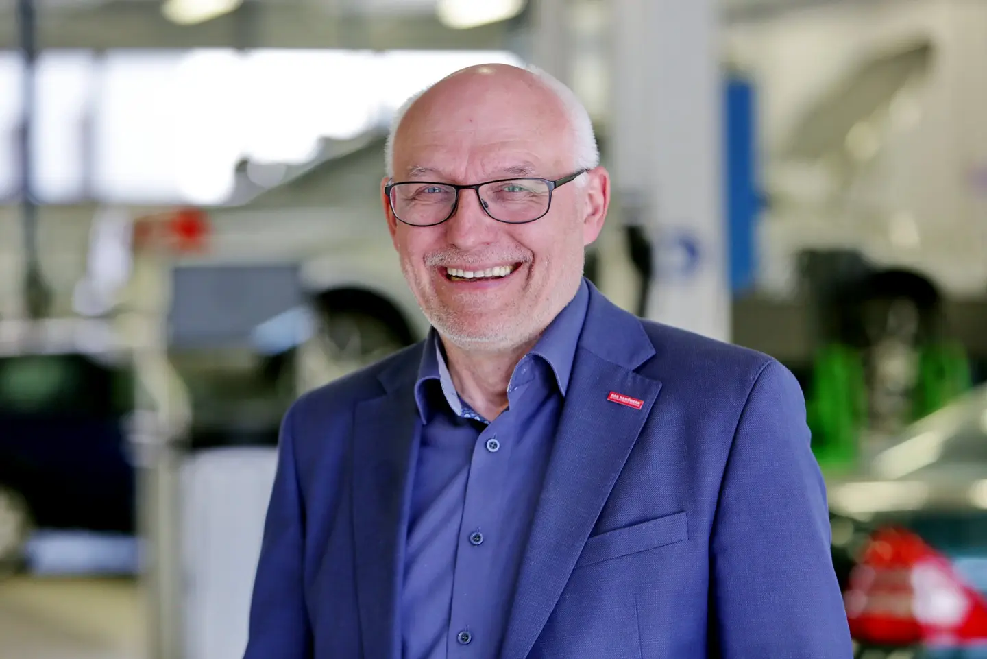 Abgebildet ist ein Mann mit kurzen grauen Haaren und ebenso kurzem grauem Bart. Er trägt ein blaues Sakko zum ebenso blauen Hemd und steht in einer KfZ-Werkstatt.