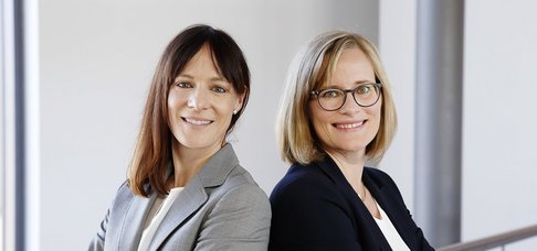 Zwei Frauen stehen Rücken an Rücken und blicken in die Kamera.