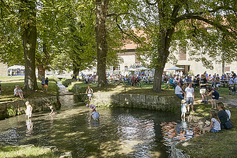Veranstaltungen auf dem Rittergut sind schon jetzt beliebt: Man sieht Menschen an Tischen und am Bachlauf im Park des Ritterguts.