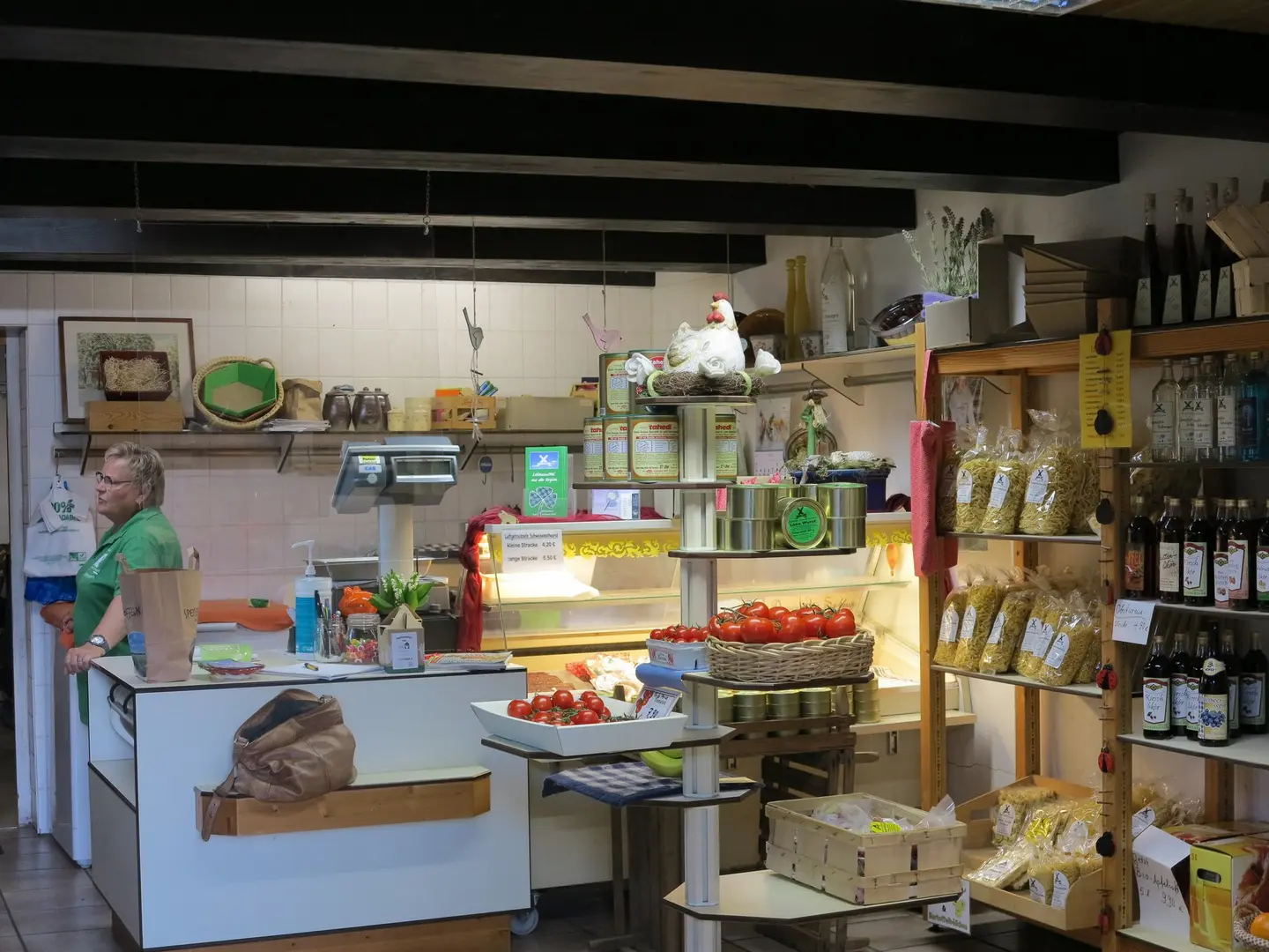 Die Aufnahme zeigt das Innere eines Hofladens voller Regale mit Nudeln, Bier, Tomaten und allerlei anderen Waren. Am Tresen steht eine Frau in einem grünen Hemd.