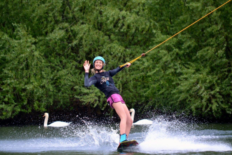 Eine Frau auf einem Wakeboard winkt in die Kamera.