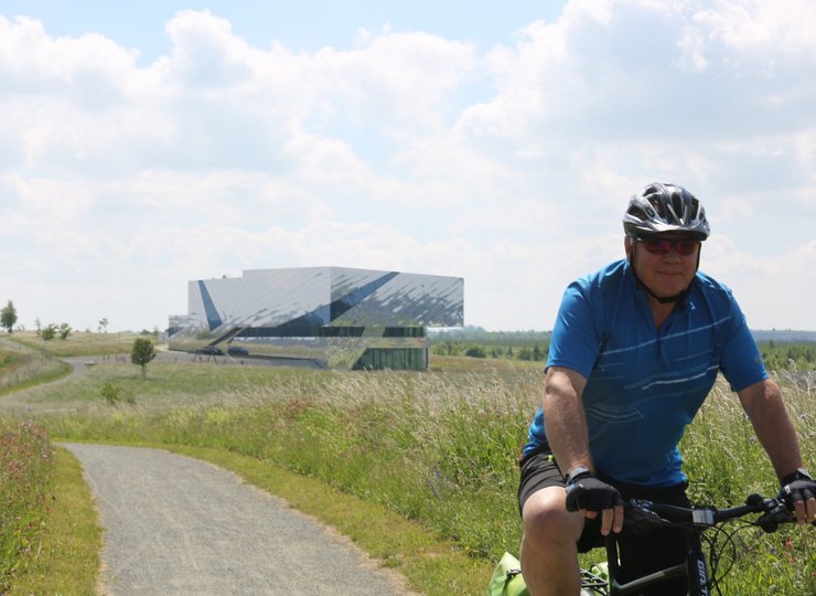 Ein Mann fährt mit seinem Rad einen Wanderweg entlang. Im Hintergrund sieht man einen modernen Glasbau.