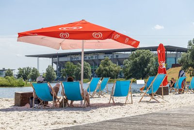 Man blickt auf die Außenberich eines Lokals am Allersee. Es stehen Liegestühle und Sonnenschirme am Stand des Allersees. Im Hintergrunnd ist die Volkswagenarena zu erkennen.
