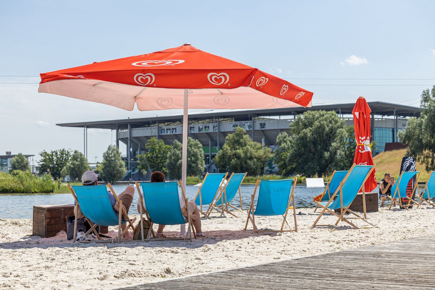 Man blickt auf die Außenberich eines Lokals am Allersee. Es stehen Liegestühle und Sonnenschirme am Stand des Allersees. Im Hintergrunnd ist die Volkswagenarena zu erkennen.