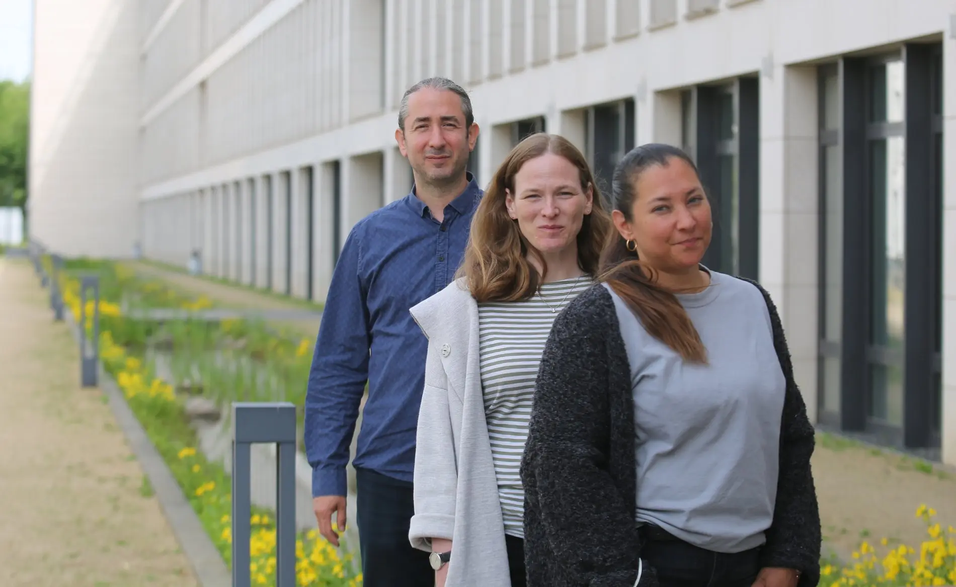 Drei Personen stehen in einer Reihe voreinander im Bild. Ein Mann steht hinten und vor ihm zwei Frauen.