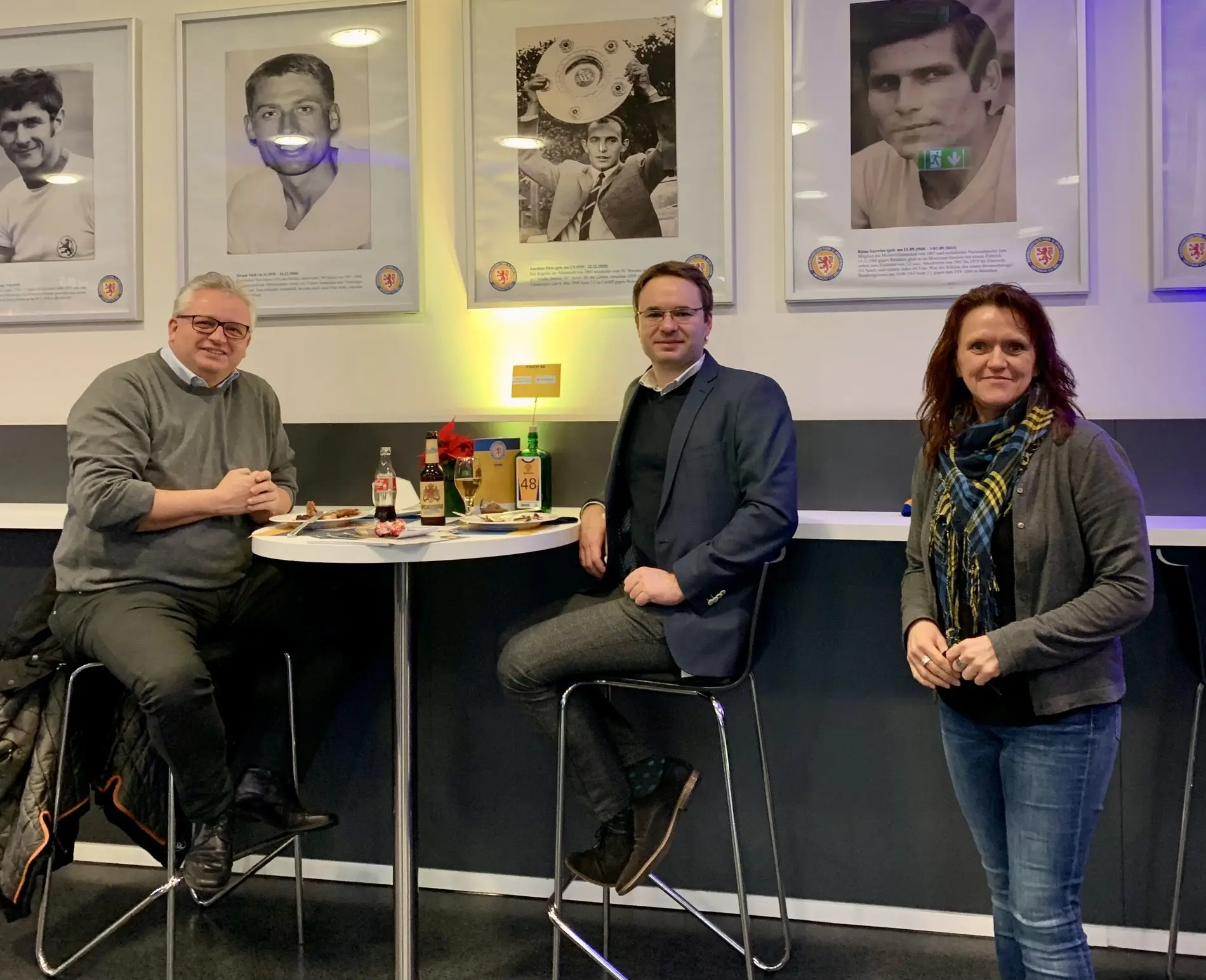 Drei Personen sitzen vor einer Wand, an der Portraitfotografien hängen.