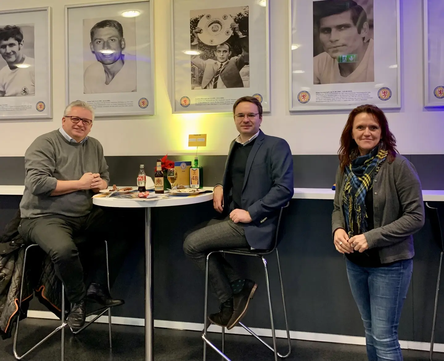 Drei Personen sitzen vor einer Wand, an der Portraitfotografien hängen.