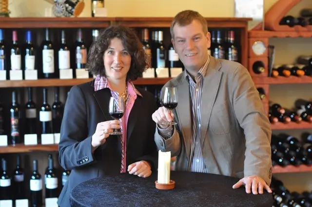Ein Ehepaar mit jeweils einem Glas Wein in der Hand, vor dem Hintergrund eines Weinregals. 