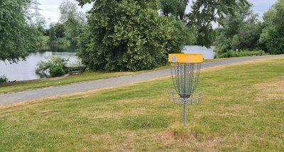 Man blickt auf einen Discgolfkorb in einer Parkanlage. Im Hintergrund liegt ein See.