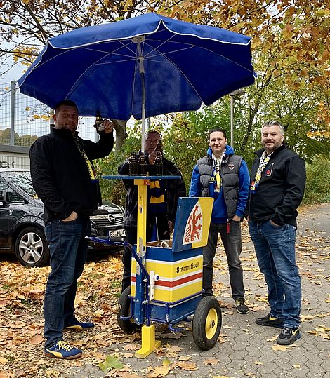 Vier Männer unter einem blauen Schirm in Eintracht-Montur.