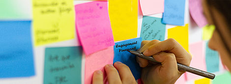 Ein Kind beschreibt einen blauen Zettel an einer bunten Pinnwand.