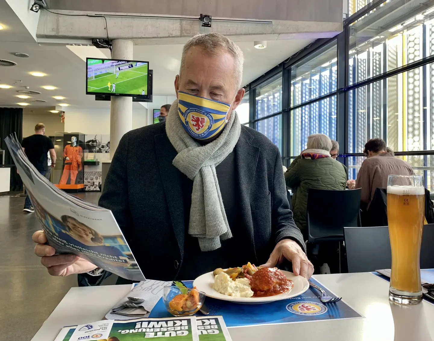 Ein Mann mit Mundschutz liest im sitzen Zeitung, vor ihm ein Mittagsgericht und ein Glas Bier. 