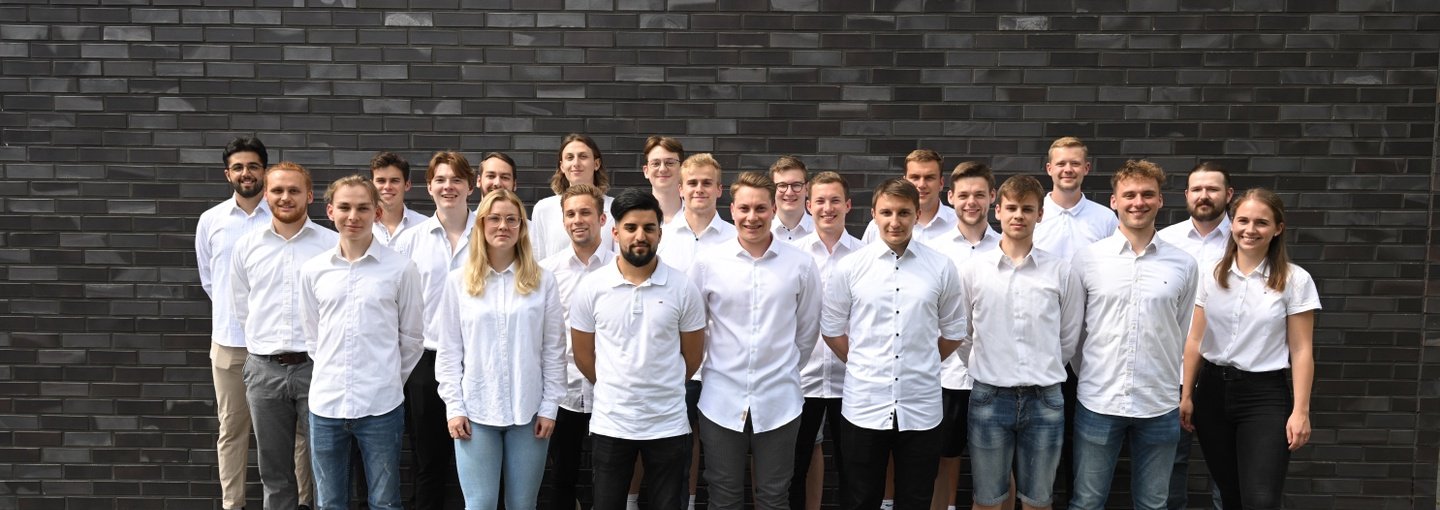 Eine Gruppe von zwanzig Studenten in weißen T-Shirts steht vor einer grauen Mauerwand.