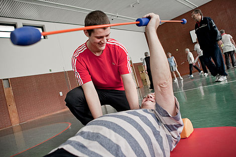 Ein junger Mann unterstützt einen älteren Menschen bei einer Sportübung. 