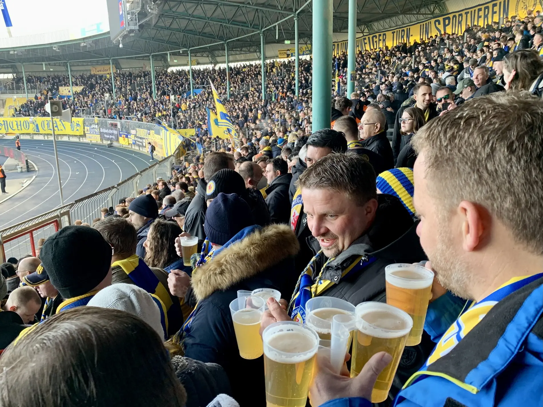 Eine volle Fankurve, im Vordergrund verteilt ein Mann Bierbecher.