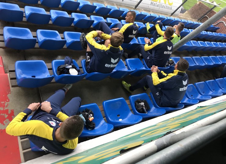 Männer in Trainingsanzügen sitzen auf blauen Sitzschalen.