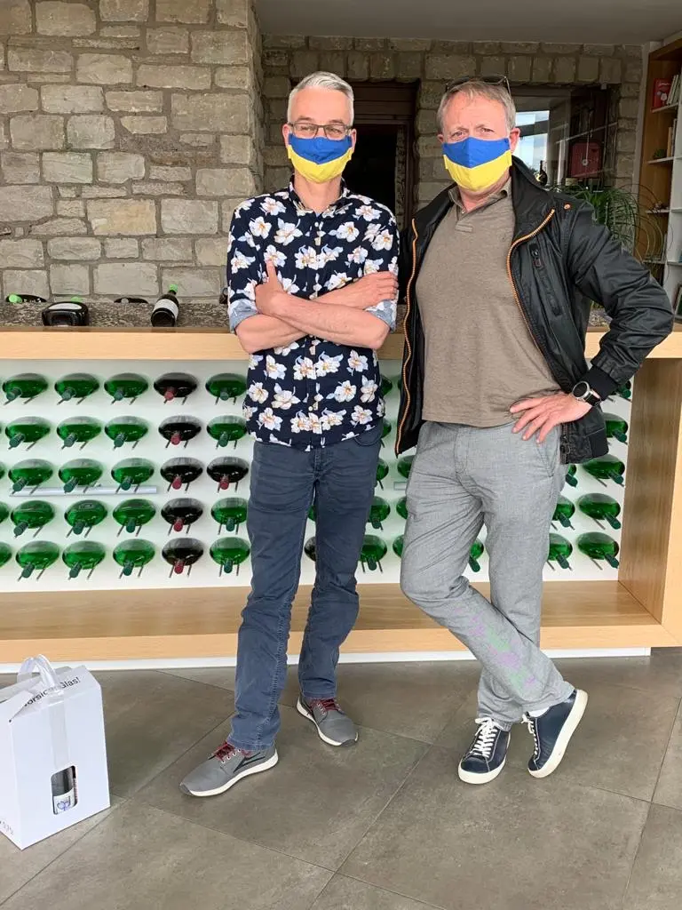 Zwei Männer mit blaugelben Mundschutzen stehen vor einem Weinregal.