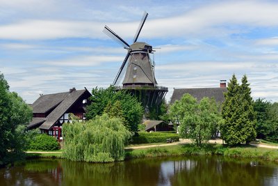 Man blickt aud einiger Entfernung auf die Sanssouci-Mühle im internationalen Mühlenmuseum Gifhorn. Neben der Mühle stehen Fachwerkhäuser. Die Mühle liegt an einem Teich.