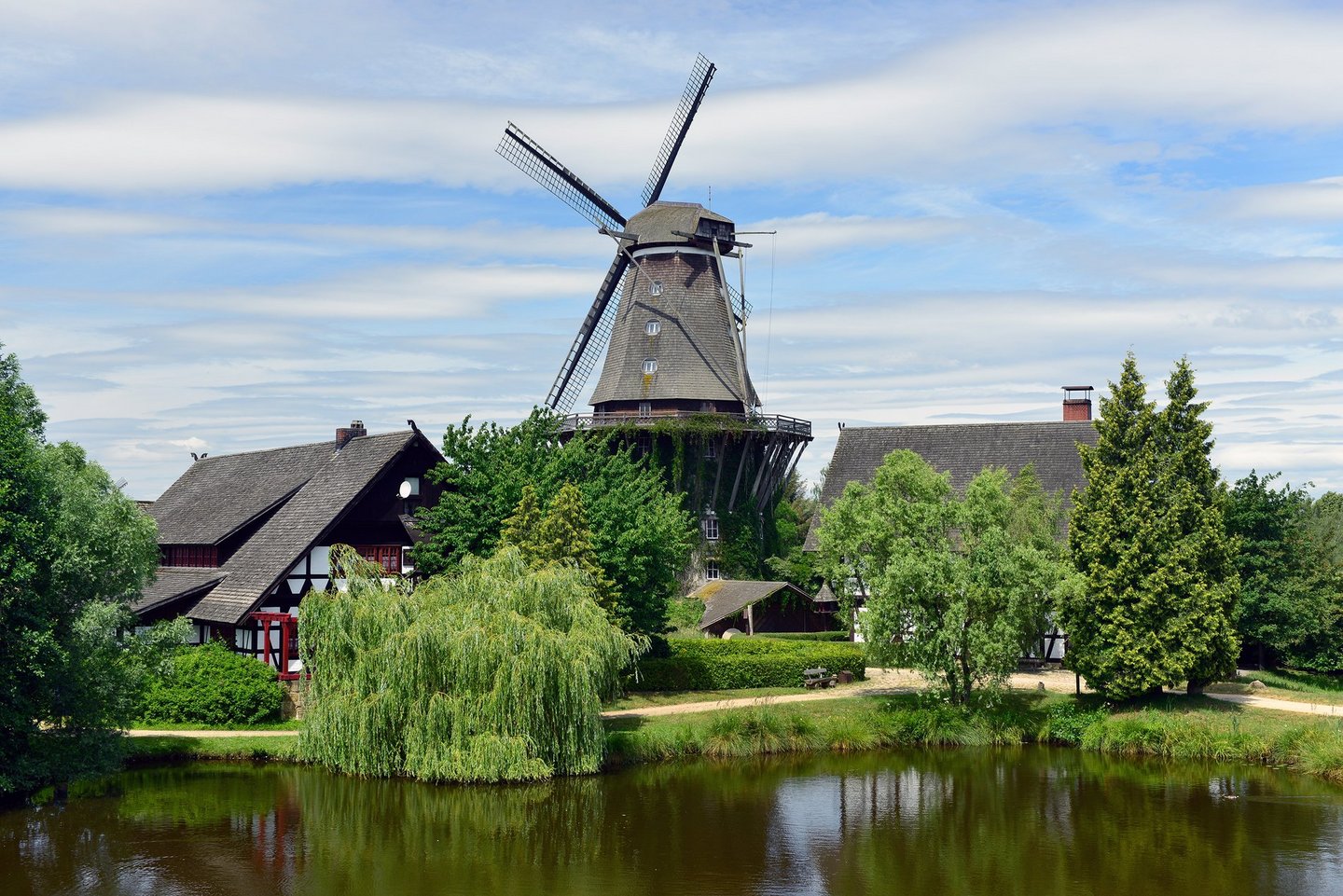 Man blickt aud einiger Entfernung auf die Sanssouci-Mühle im internationalen Mühlenmuseum Gifhorn. Neben der Mühle stehen Fachwerkhäuser. Die Mühle liegt an einem Teich.