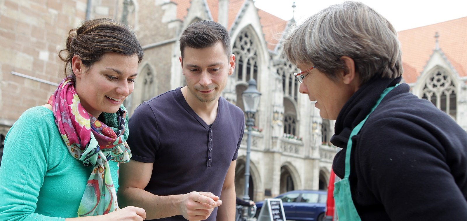Ein Mann und eine Frau stehen bewundernd einer Marktfrau gegenüber und bestaunen den von ihr angebotenen Spargel.