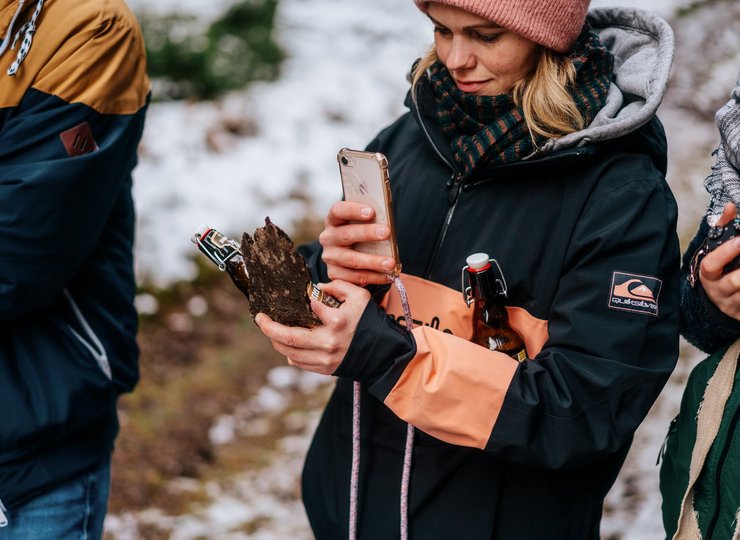 Eine Frau in winterlicher kleidung fotografiert mit ihrem Handy ein Stück Baumrinde. Unter dem Arm hat sie eine Bierflasche eingeklemmt. 
