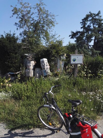 Man blickt auf die Gabelung zweier Feldwege. Auf der begrünten Fläche sind Holzskulturen menschlicher Gesichter zu erkennen. Rechts daneben steht ein Hinweisschild.