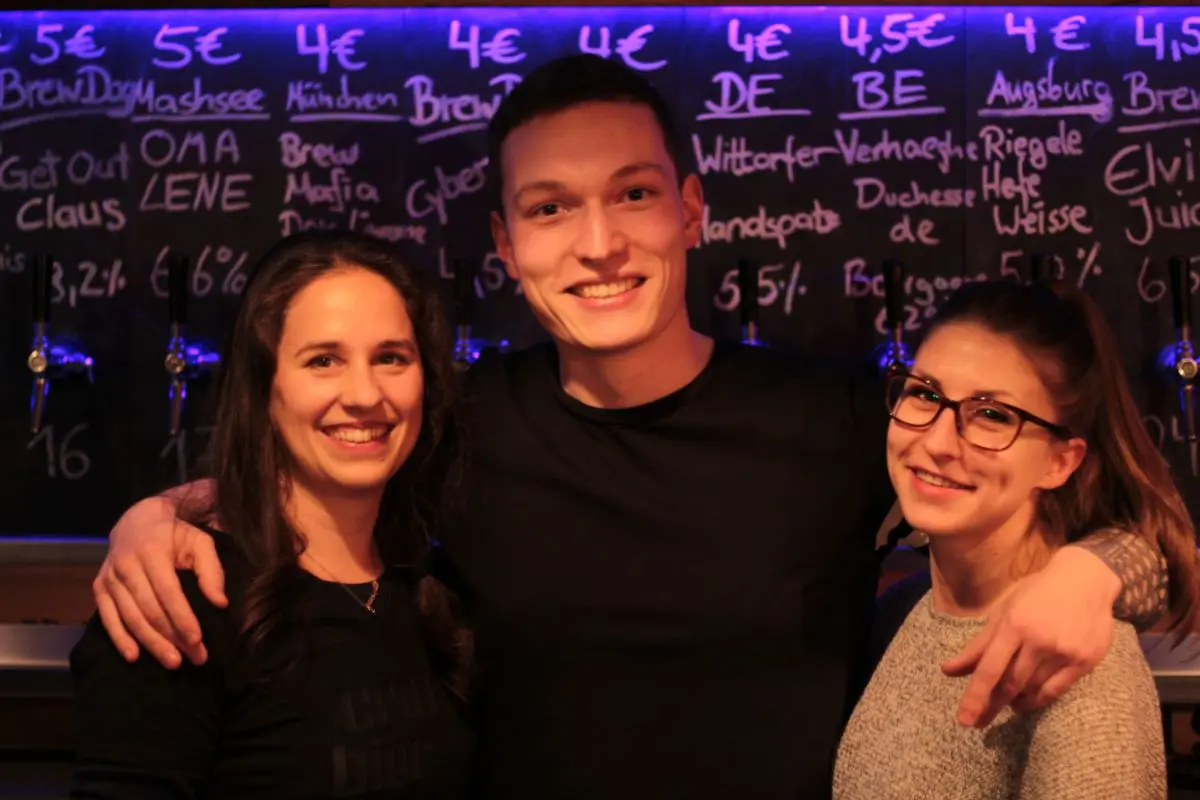 Ein junger Mann hält zwei junge Frauen in den Armen, im Hintergrund eine Thekeneinrichtung.